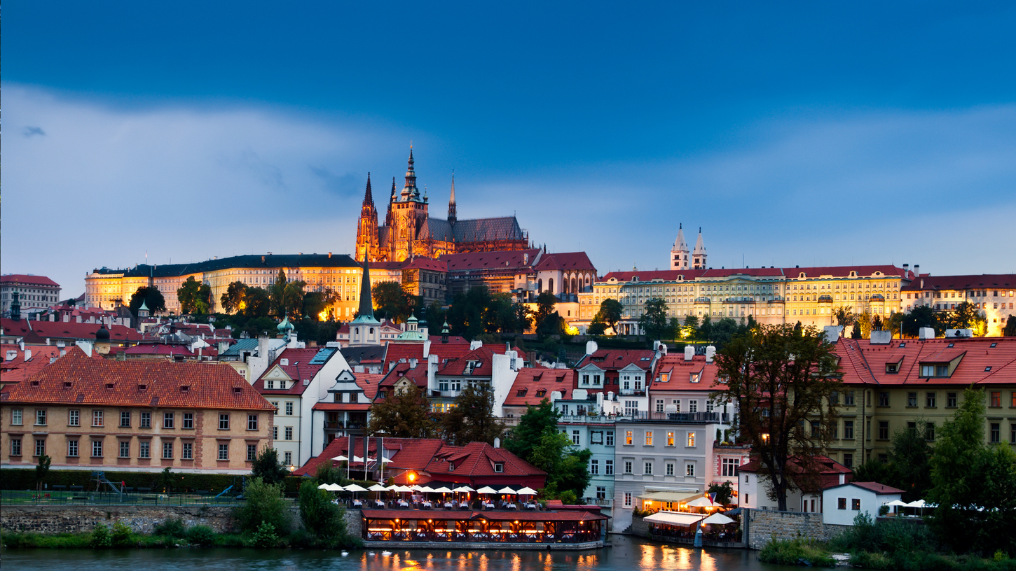 A breathtaking view of the iconic Prague Castle, bathed in the soft, golden hues of twilight. This enchanting scene showcases the majestic castle perched atop its hill, surrounded by a picturesque ensemble of historic Prague houses. The castle's lights create a captivating contrast against the gradually darkening sky, forming a truly magical sight in the heart of the Czech capital.
