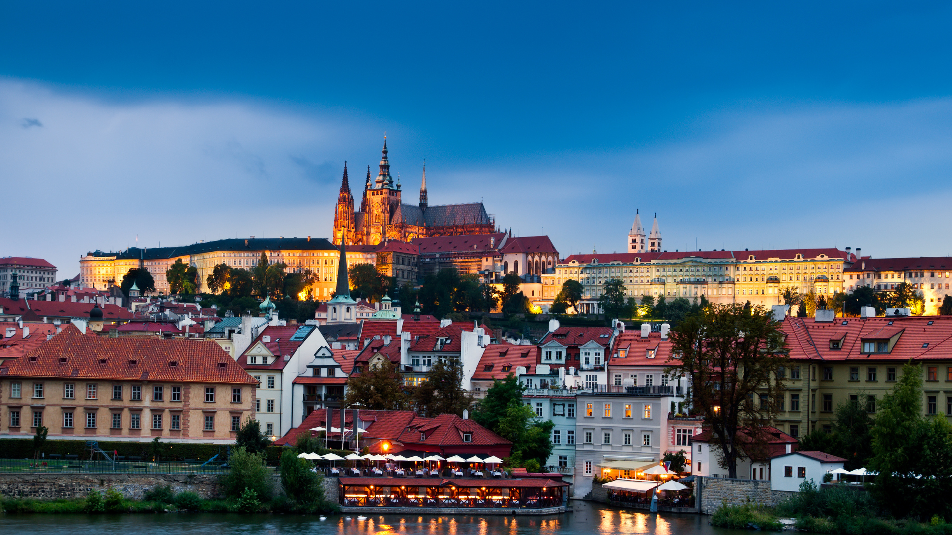 A breathtaking view of the iconic Prague Castle, bathed in the soft, golden hues of twilight. This enchanting scene showcases the majestic castle perched atop its hill, surrounded by a picturesque ensemble of historic Prague houses. The castle's lights create a captivating contrast against the gradually darkening sky, forming a truly magical sight in the heart of the Czech capital. 