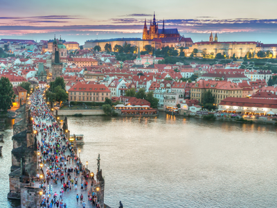  Prague Castle Illuminated at Dusk: This enchanting image captures the Prague Castle aglow with the warm, welcoming lights of evening. It stands prominently amidst a picturesque scene of historic Prague houses, and the Charles Bridge, adorned with a bustling crowd, is seen nearby. The combination of architectural splendor and a lively atmosphere makes this a quintessential Prague experience, perfect for exploring the city's rich heritage and vibrant culture.